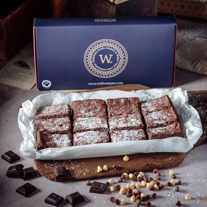 A boxed selection of chocolate chip brownies dusted with powdered sugar, displayed on parchment paper with scattered chocolate pieces and chocolate chips in front, and a blue Wilfreds branded box behind them.