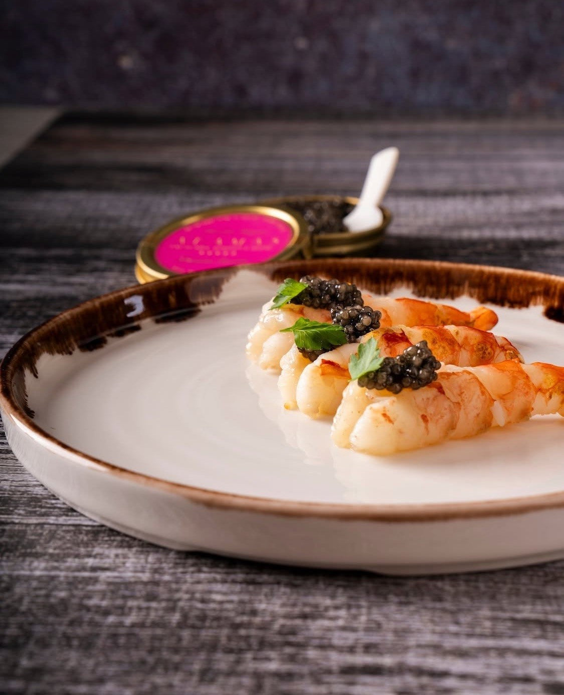 Prawns topped with caviar, presented on a white plate, with an open tin of pink label Adamas caviar in the background and a mother of pearl spoon inside.