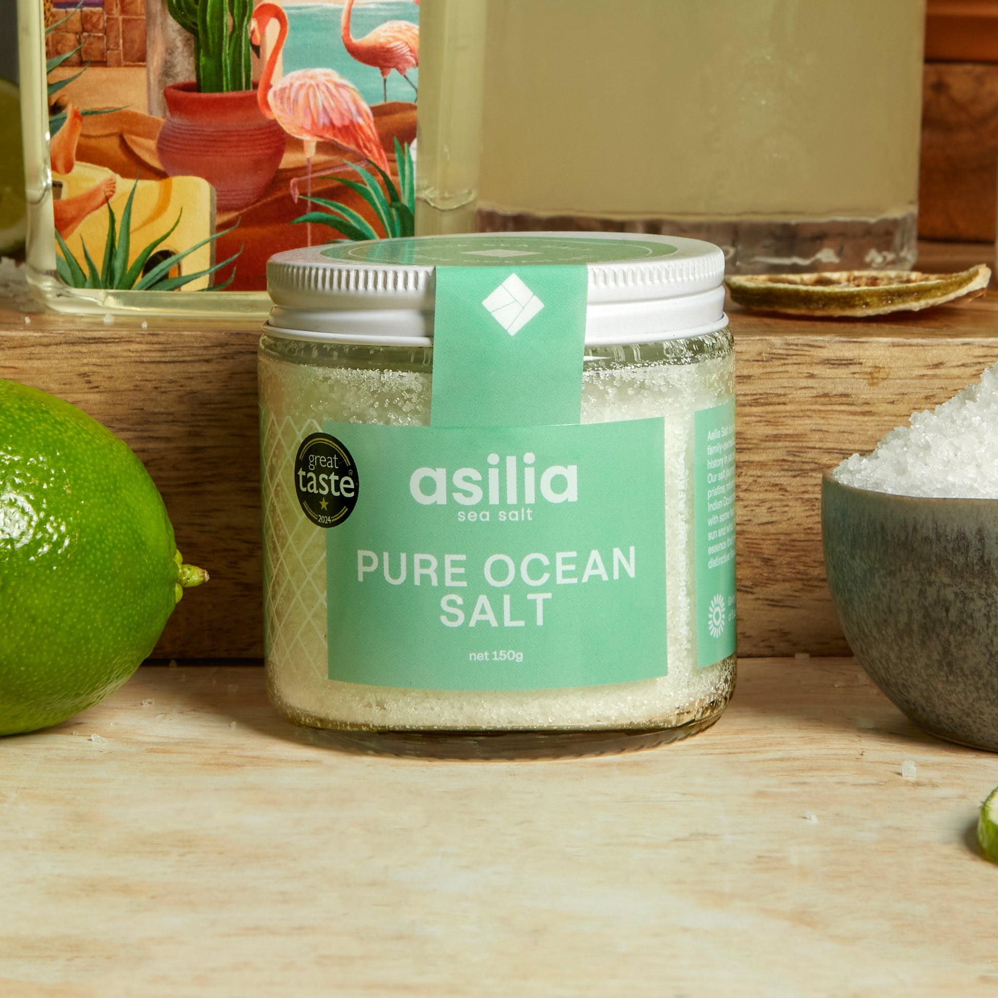 Jar of asilia pure ocean salt on a wooden surface with a lime and decorative items in the background.