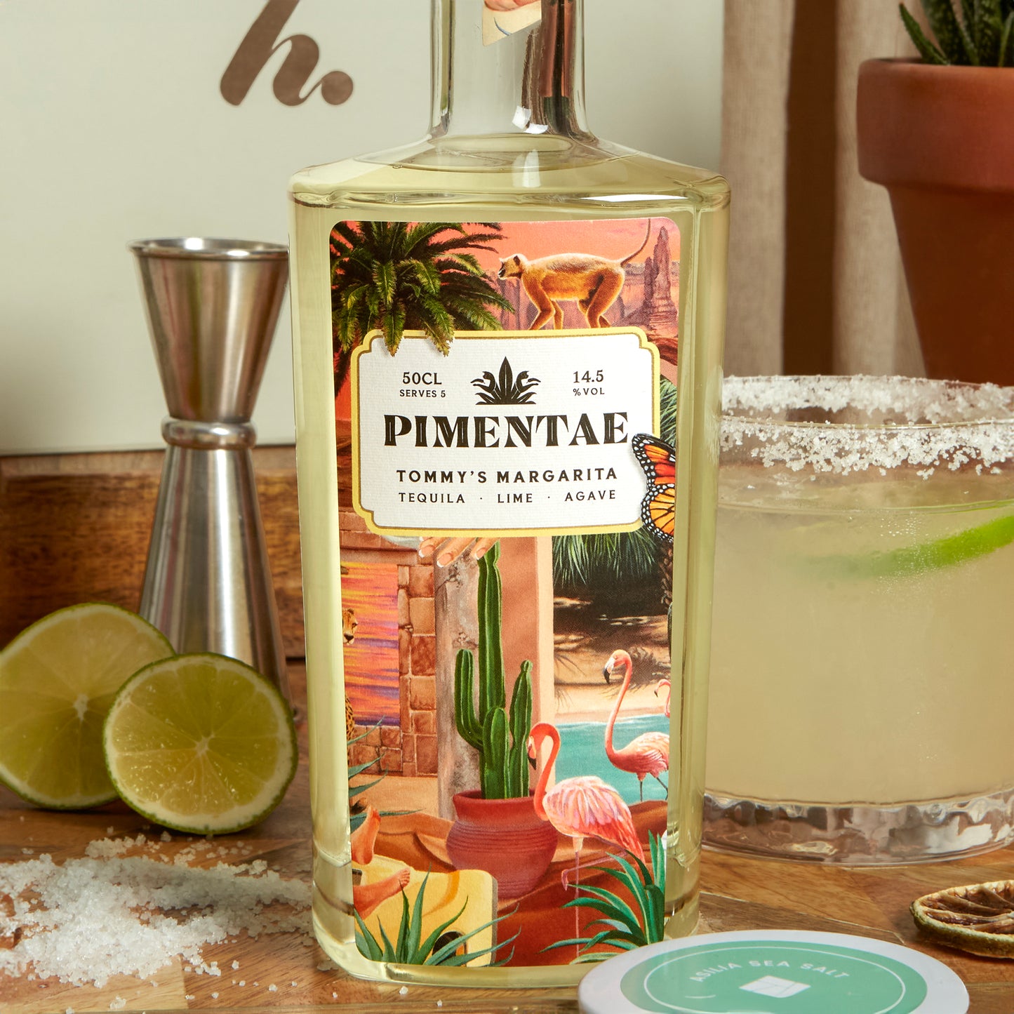 Pimentae Tommy's Margarita bottle with a scenic label, surrounded by limes and a cocktail glass.