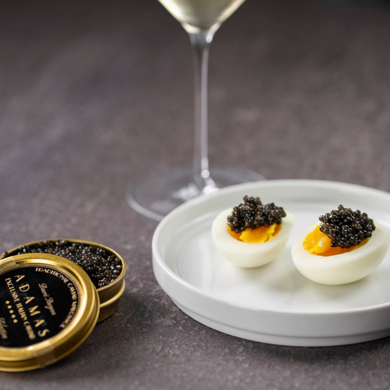 Two halves of a devilled egg topped with caviar on a small white plate, with an open round tin of Adamas Prestige Selection caviar and a wine glass stem visible in the background on a dark slate surface.