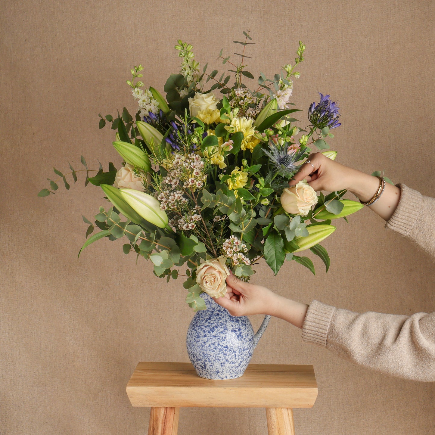 Fresh, seasonal flowers in a large bouquet arrangement, displayed in a blue and white jug vase, styled on a wooden stool with a person's hands adjusting the flowers, against a neutral beige background.
