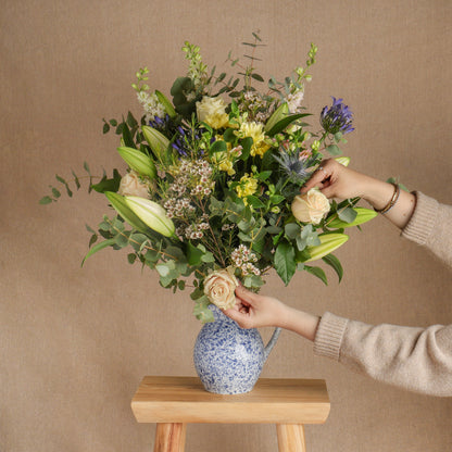 Fresh, seasonal flowers in a large bouquet arrangement, displayed in a blue and white jug vase, styled on a wooden stool with a person's hands adjusting the flowers, against a neutral beige background.