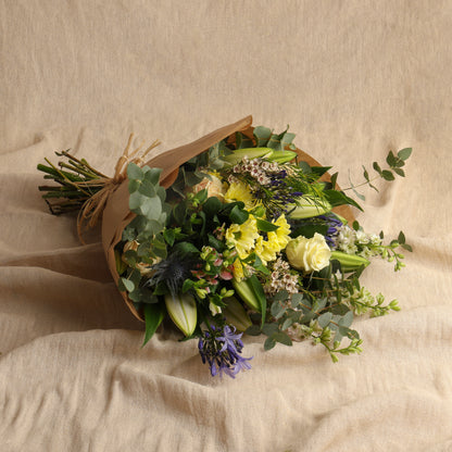 Large seasonal flower bouquet delivered hand-tied and wrapped in brown gift paper, displayed on draped beige fabric.
