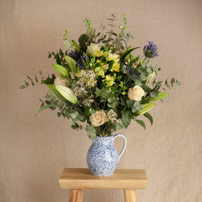 Large seasonal flower bouquet arranged in a blue and white jug vase, displayed on a small wooden stool against a neutral beige backdrop.