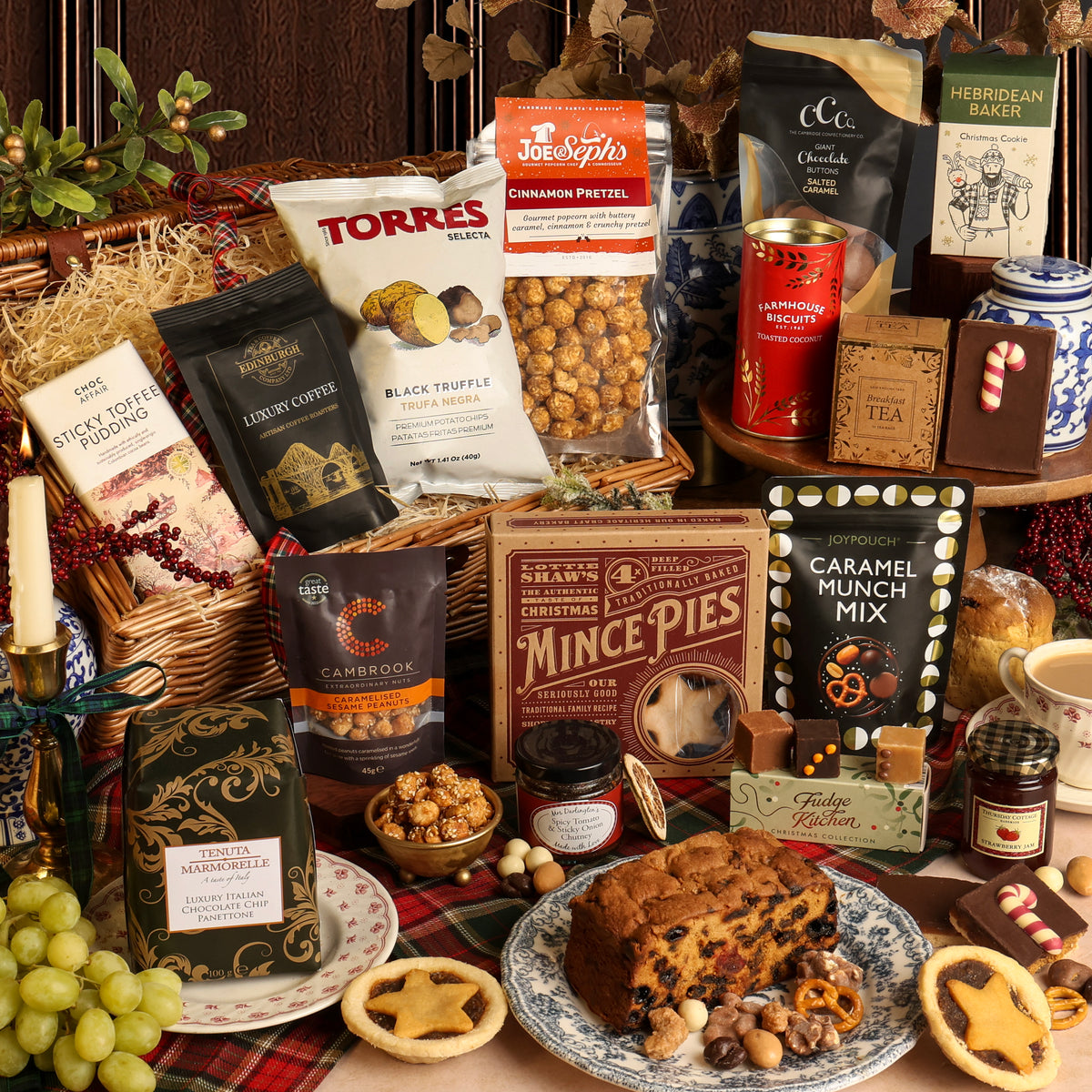 The Magic of Christmas gift hamper filled with assorted snacks, tea, coffee, and baked goods arranged on a table with a plaid runner. Visible items include a loaf of fruit cake, mince pies, nuts, and chocolate.