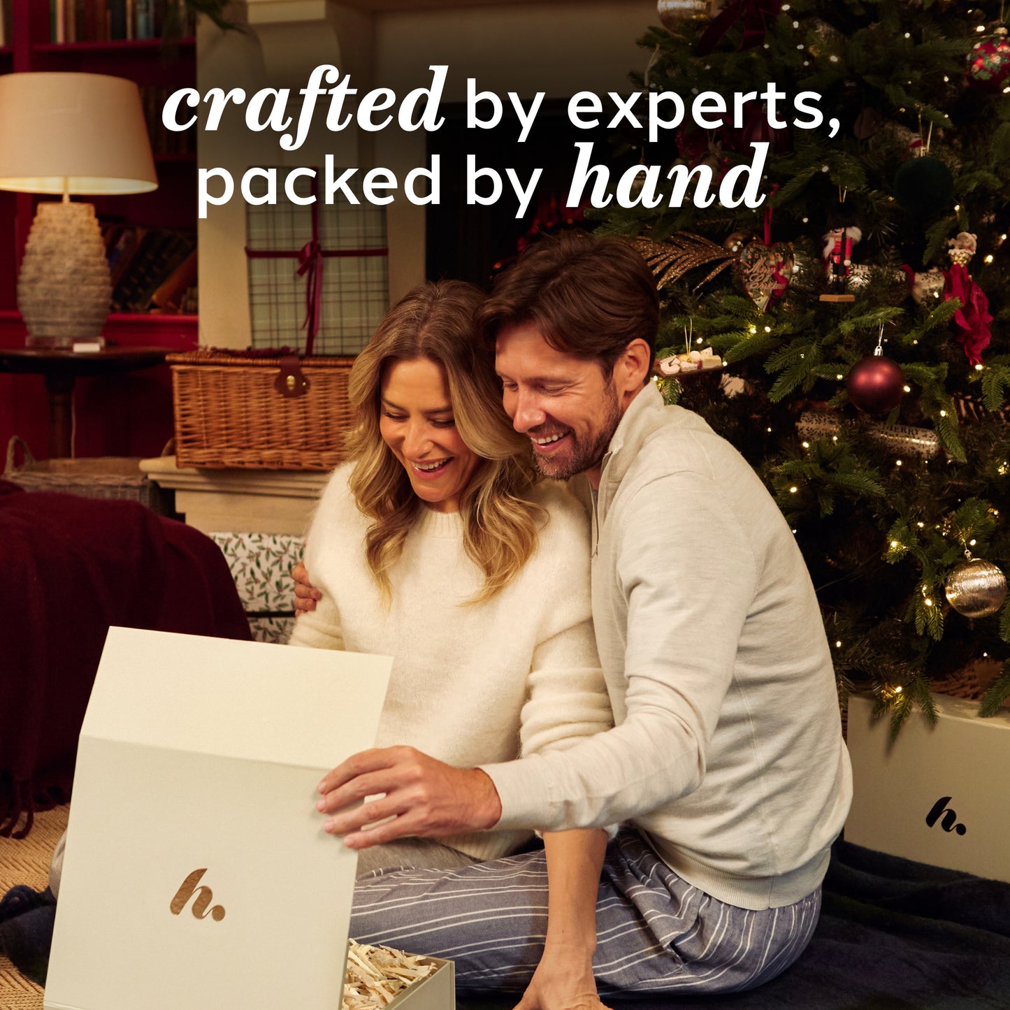 Couple opening a gift in front of a Christmas tree with text 'crafted by experts, packed by hand'.