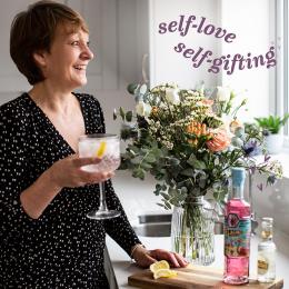 Image of woman holding a gin glass looking out of a window with flowers, gin and tonic in front of her, with text 'self-love self gifting'