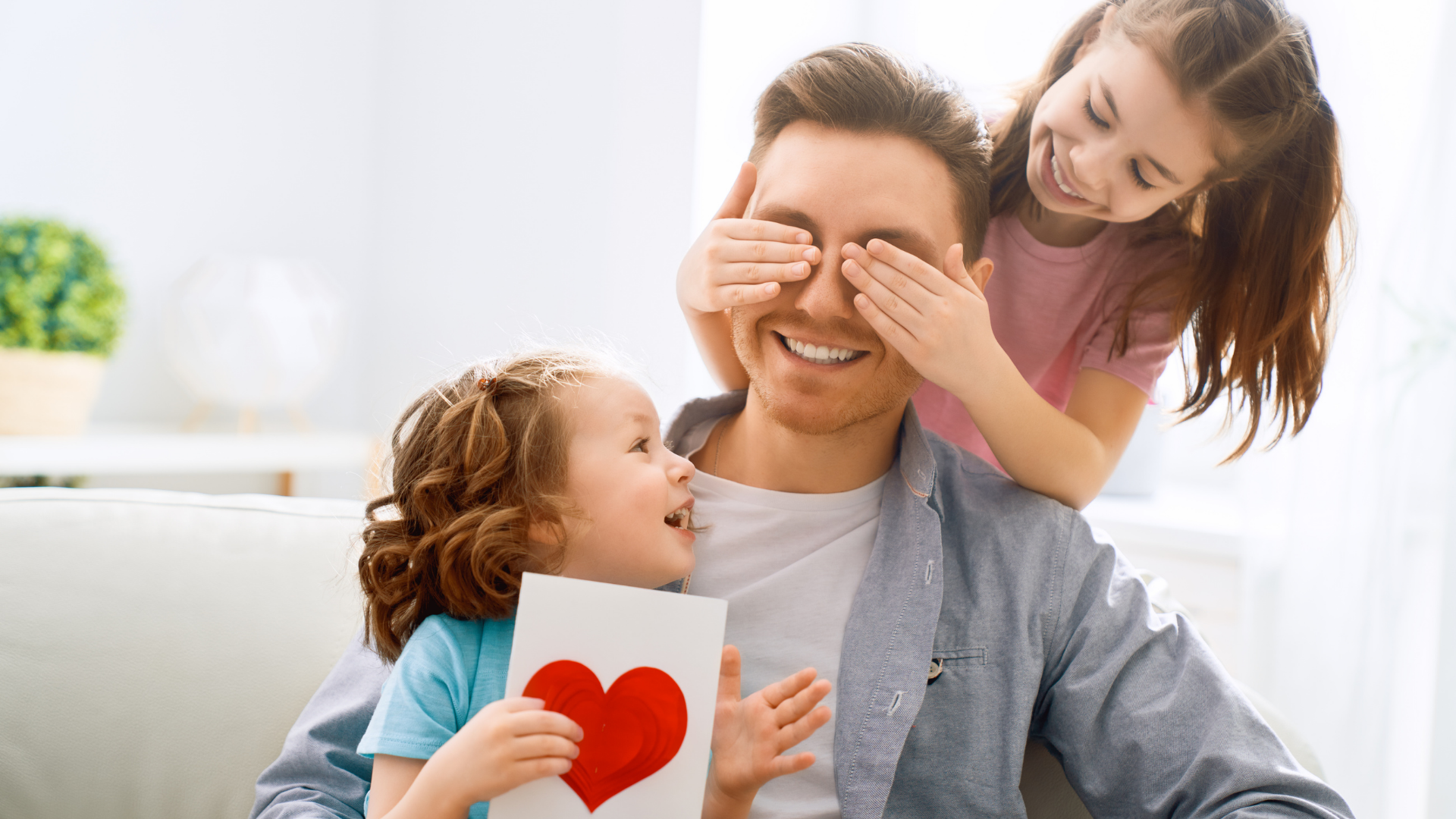 One daughter sat on dad's lap giving him a heart card whilst other daughter covers his eyes to surprise him on Father's Day