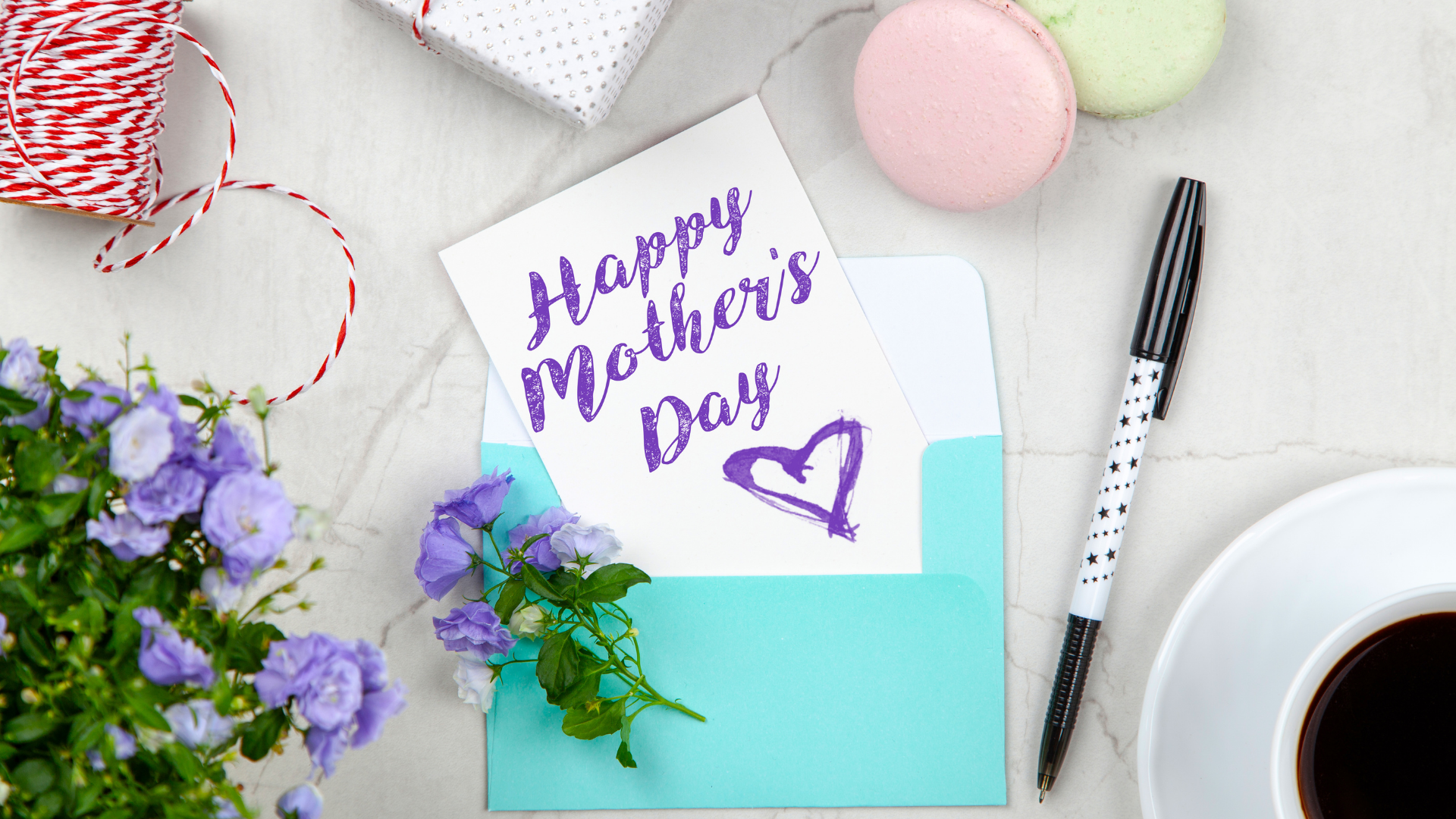 Happy mother's day written on notecard, surrounded by flowers, string, a pen and macarons