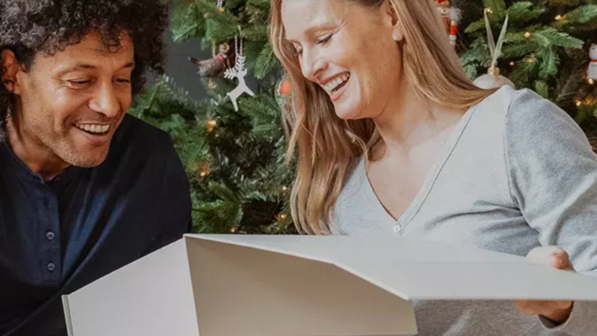 Couple looking at each other opening a hampers.com Christmas gift box with Christmas tree in the background