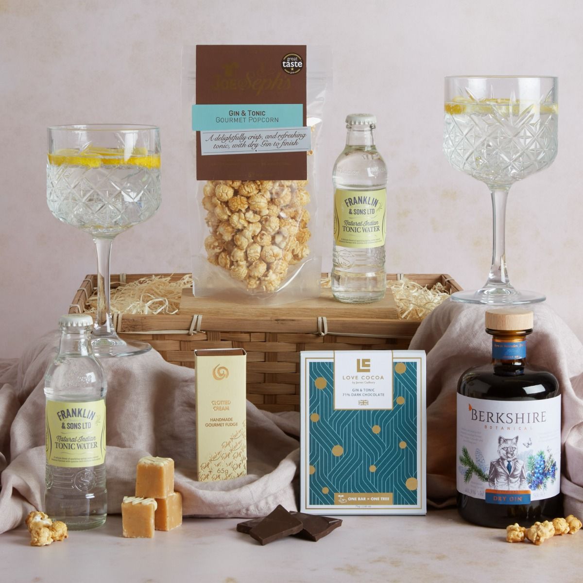 The Luxury Gin Hamper with contents on display and two glasses on gin and tonic with gift basket