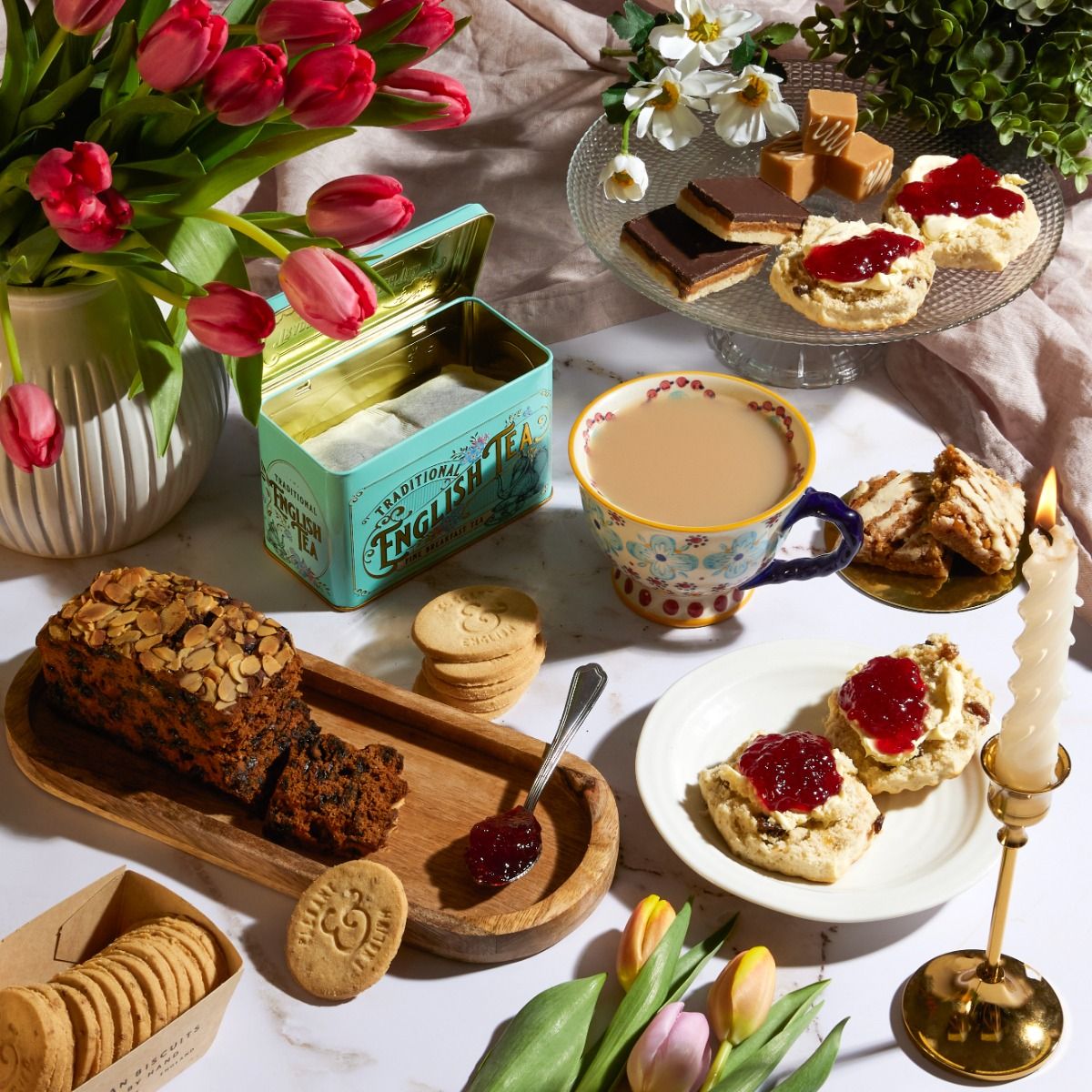 A cup of tea surrounded by cake, flowers and a tin of tea bags